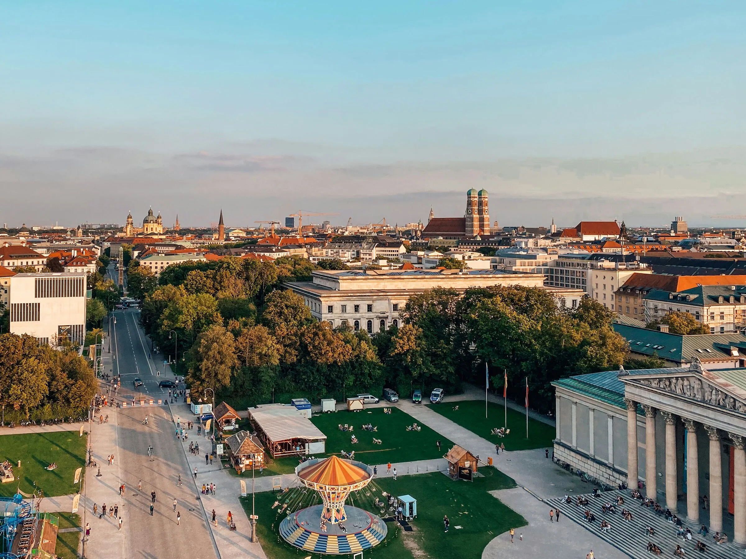 Blick von oben auf München am Abend: ein Platz mit Kettenkarussell, dahinter die Dächer der Innenstadt und die Türme der Frauenkirche